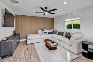 A modern living room with a sectional sofa, a wooden accent wall, a ceiling fan, and a window with a view of greenery. - Scottsdale Burro! Resort Pool, Casita & Putting (Scottsdale)