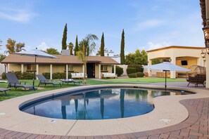 Ein Schwimmbecken mit Liegestühlen und Sonnenschirmen, umgeben von einem angelegten Garten und einem Gebäude mit beige gefärbter Fassade.