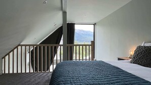 Ein Schlafzimmer mit Bett, Nachttisch, Lampe, Fenster mit Bergblick und Balkon mit Geländer.