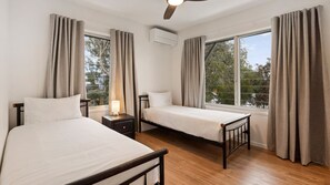 Ein Schlafzimmer mit zwei Betten, einem Nachttisch mit Lampe, einer Klimaanlage und großen Fenstern mit Vorhängen.