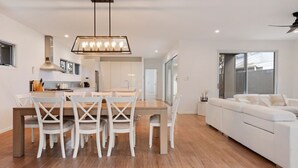 Een moderne keuken met witte kasten, een eethoek met een houten tafel en witte stoelen, en een witte bank in een woonruimte met grote ramen.