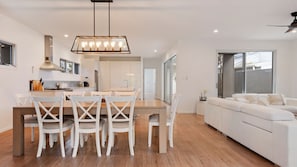 Eine moderne Küche mit weißen Schränken, ein Essbereich mit Holztisch und weißen Stühlen sowie ein weißes Sofa in einem Wohnbereich mit großen Fenstern.