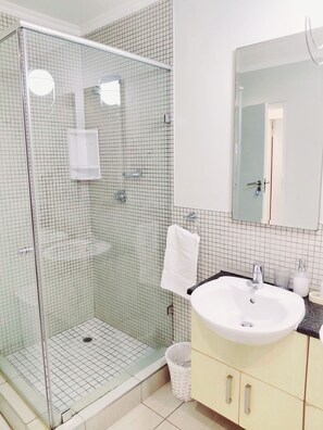 Uma casa de banho com cabine de duche em vidro, uma sanita branca com espelho em cima e um contentor de lixo branco.
