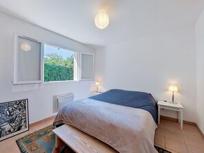 A bedroom with a bed, two bedside lamps, a window, a radiator, and a framed picture on the wall. - Les Mouettes by Interhome (Soustons)