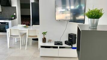A modern living space with a flat-screen TV mounted on the wall, a dining area with white chairs, a kitchen with a black stove, and a potted plant on the sideboard.