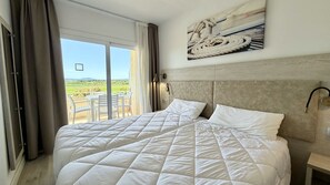 Ein großes Bett mit weißer, gesteppter Decke, ein Kopfteil mit heller Holzoptik, eine Wandleuchte und Blick auf eine Terrasse mit Gartenmöbeln.