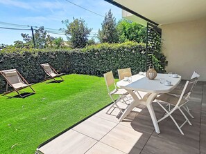 Eine Terrasse mit Esstisch und Stühlen, ein kleiner Garten mit Rasen und einer Hecke und Blick auf den Himmel.