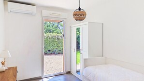 Ein Schlafzimmer mit Bett, Nachttisch, Lampe, Deckenventilator und Blick auf einen Garten durch eine Schiebetür.