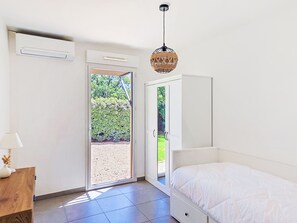 Ein Schlafzimmer mit Bett, Nachttisch, Lampe, Deckenventilator und Blick auf einen Garten durch eine Schiebetür.