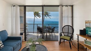 Ein Balkon mit Blick auf den Ozean, ein Esstisch, Stühle, eine Couch und ein Fernseher.