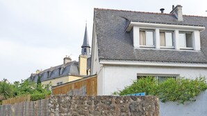 Un mur de pierre, un bâtiment blanc avec un toit gris et, au loin, le clocher d’une église.