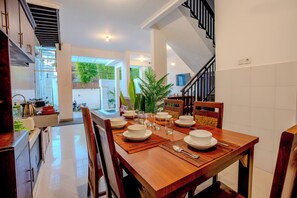 A dining area with a wooden table set for four, surrounded by chairs, with a staircase and a view of greenery outside. - Cedil Private 3BR Pool Villa Seminyak (Kabupaten Badung)