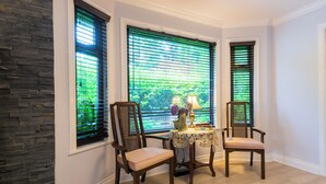 A corner room with three windows, a table with a vase and books, and two chairs.