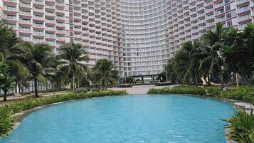 Ein Swimmingpool mit zwei Personen im Wasser, umgeben von Palmen und einem großen Hotel im Hintergrund.