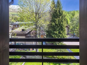 Blick durch ein Fenster mit Metallgitter auf eine grüne Landschaft mit Bäumen, ein Gebäude mit Satteldach und ein Berg im Hintergrund.
