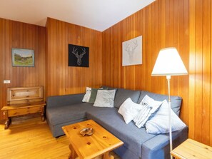 Een kamer met houten lambrisering, een grijze bank, een houten tafel, een bankje en een lamp.