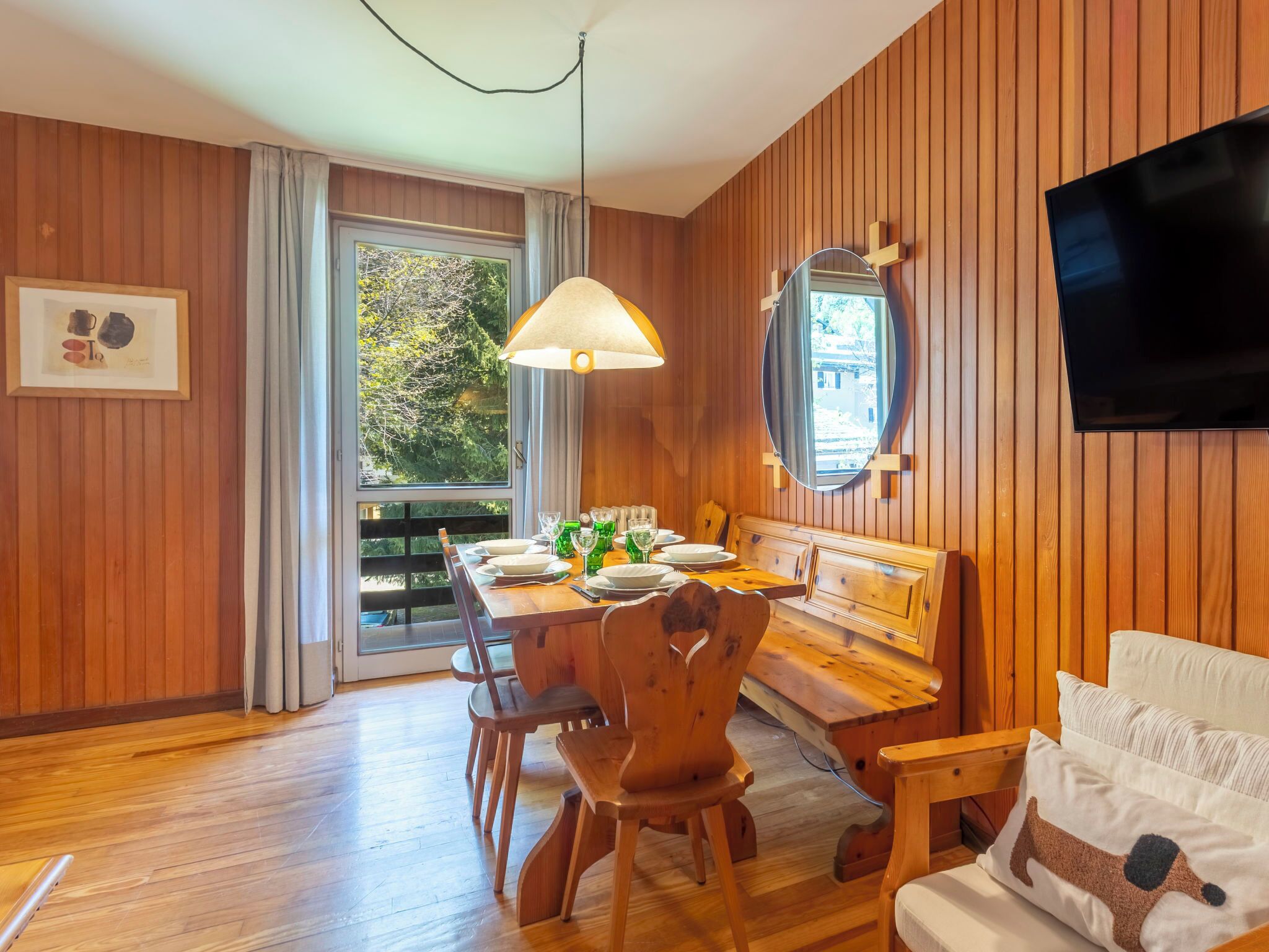 Une pièce aux murs lambrissés, avec une table à manger dressée pour quatre personnes, un canapé, un téléviseur fixé au mur et une fenêtre donnant sur la forêt.