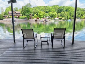 Une terrasse avec deux chaises et une petite table, donnant sur un lac paisible où l’on aperçoit des bateaux, et sur une zone boisée en arrière-plan.
