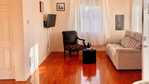 Ein Wohnzimmer mit Holzboden, einem schwarzen Sessel, einem beigen Sofa, einem kleinen schwarzen Tisch und einem an der Wand montierten Flachbildfernseher.