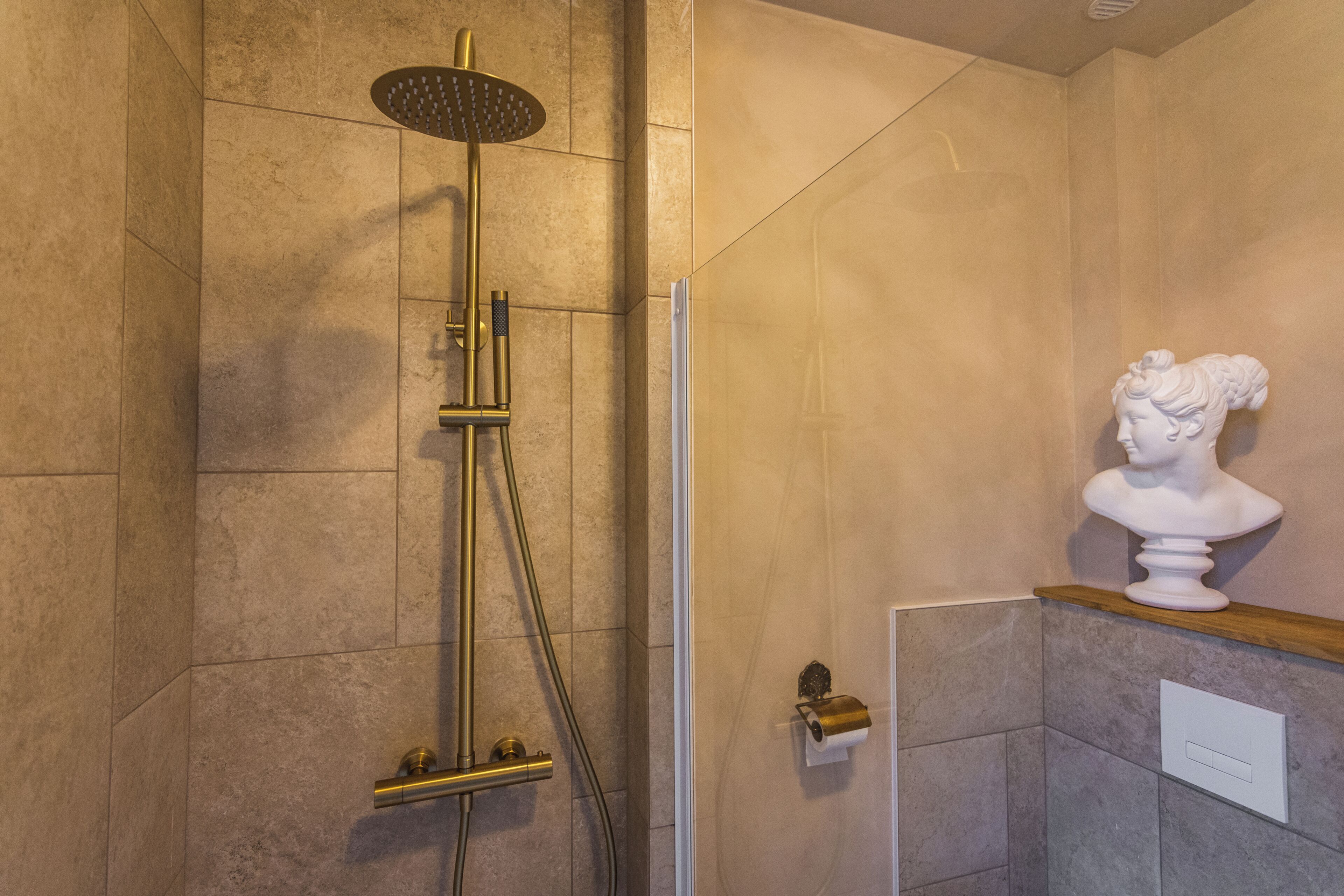 A shower with a gold showerhead and control panel, a marble bust on a wooden shelf, and a white flush toilet.