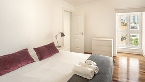 Una camera da letto con un letto, cuscini, un comodino con una lampada, una cassettiera e una finestra vista.