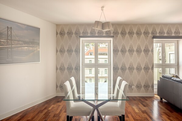 A dining area with a glass table, two chairs, a patterned wallpaper, a large window, and a view of a bridge. - Downtown Riverside V by Homing (Lisbon)