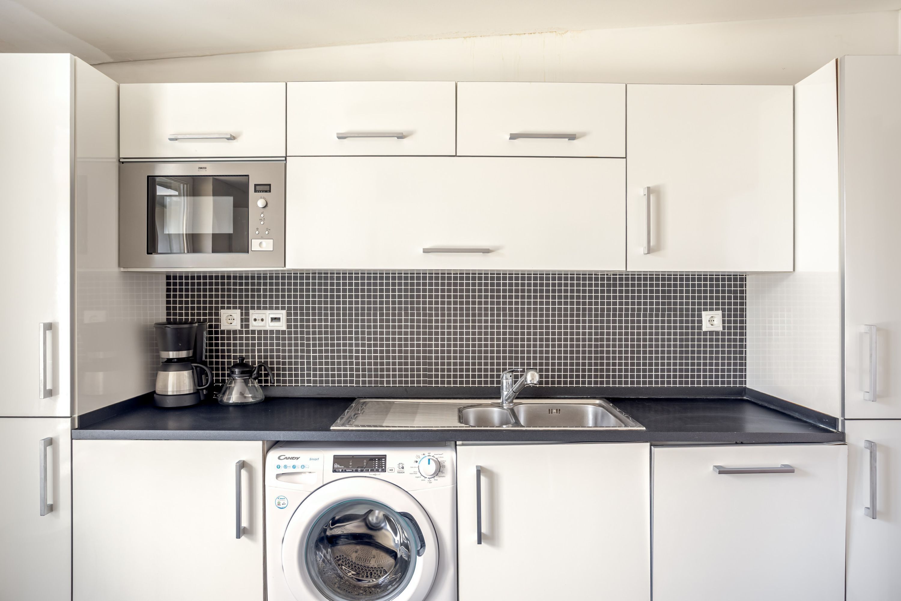 Una cocina moderna con armarios blancos, microondas, cafetera y lavadora.