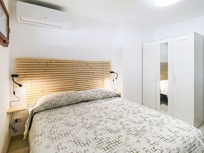 Ein Schlafzimmer mit einem Bettgestell aus Holzleisten, einer gemusterten Bettdecke, einer Nachttischlampe, einer Klimaanlage und einem Kleiderschrank.