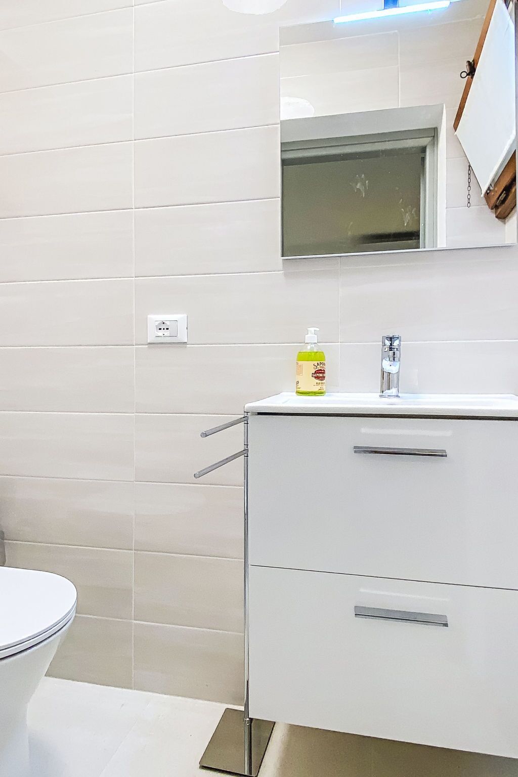 Et badeværelse med hvide flisevægge, en hvid vask med et spejl over og et toilet.