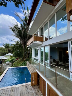 Ein modernes Haus mit Swimmingpool, großen Fenstern und einem Balkon mit Blick auf üppiges Grün.