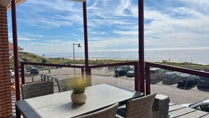 A balcony with a table and chairs, a potted plant, and a view of the beach and parking area. - Beautifully located on Nordboulevard on the 1st floor. Directly on the beach, balcony with wonderful sea view, 2 bedrooms and garage. (Egmond aan Zee)