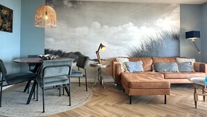 A living room with a modern sofa, a dining table with chairs, a hanging light fixture, and a mural on the wall. - Beautifully located on Nordboulevard on the 1st floor. Directly on the beach, balcony with wonderful sea view, 2 bedrooms and garage. (Egmond aan Zee)