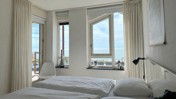 A hotel room with two beds, a desk, a chair, and large windows offering a view of the sea. - Beautifully located on Nordboulevard on the 1st floor. Directly on the beach, balcony with wonderful sea view, 2 bedrooms and garage. (Egmond aan Zee)