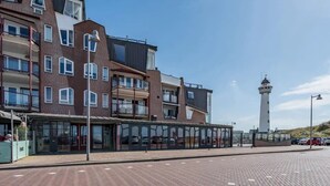 A modern building with multiple floors, a glass facade, and a lighthouse in the background. - Beautifully located on Nordboulevard on the 1st floor. Directly on the beach, balcony with wonderful sea view, 2 bedrooms and garage. (Egmond aan Zee)