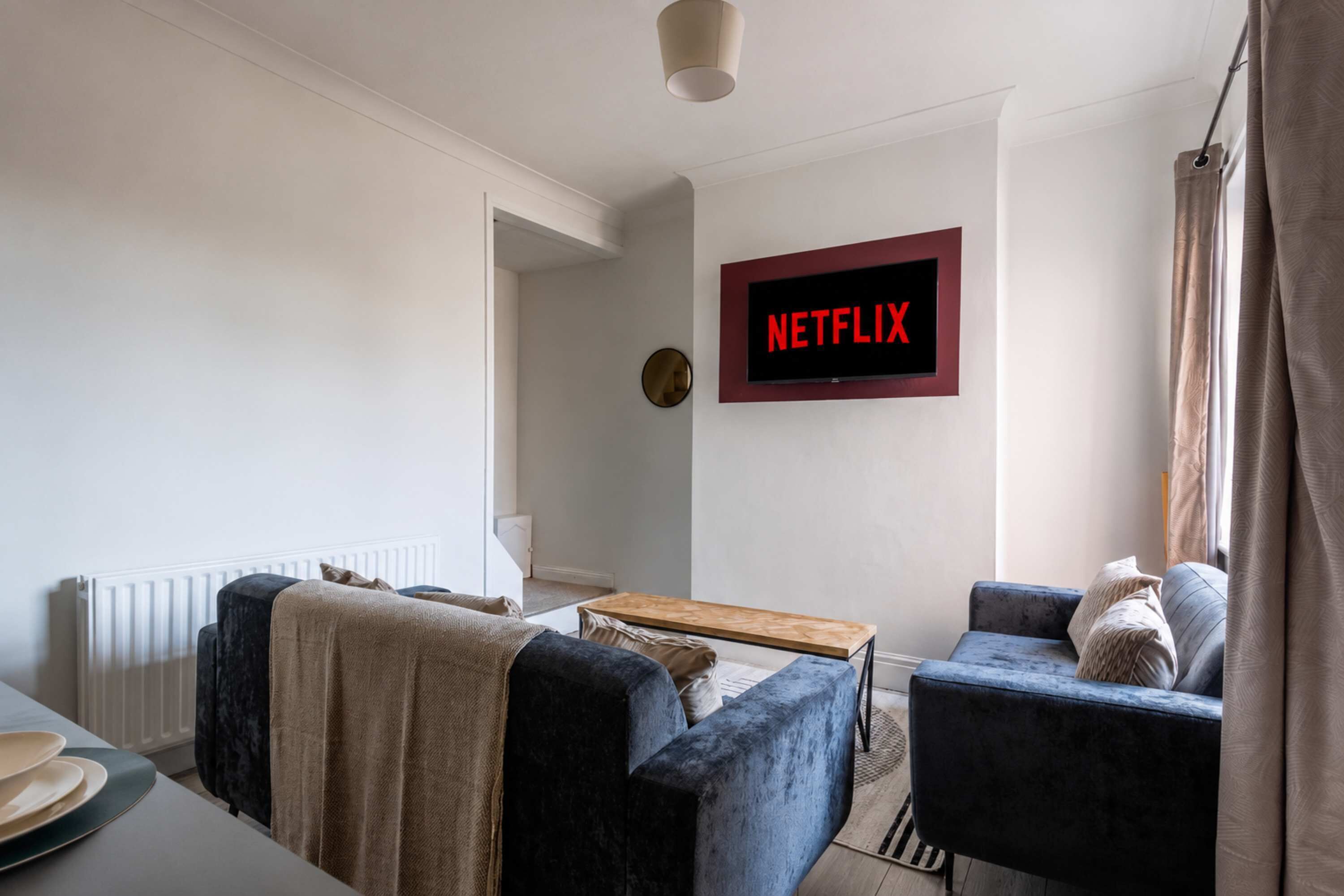 A living room with a sofa, armchair, coffee table, and a wall-mounted TV displaying 'NETFLIX'.