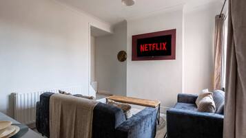 A living room with a sofa, armchair, coffee table, and a wall-mounted TV displaying 'NETFLIX'.