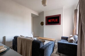 Een woonkamer met een bank, fauteuil, salontafel en een aan de muur bevestigde televisie waarop 'NETFLIX' wordt getoond.