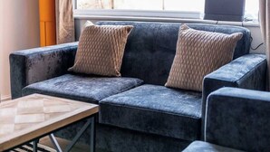 Un sofá azul oscuro con dos almohadones trenzados, una mesa de centro de madera y un ordenador portátil en un alféizar.