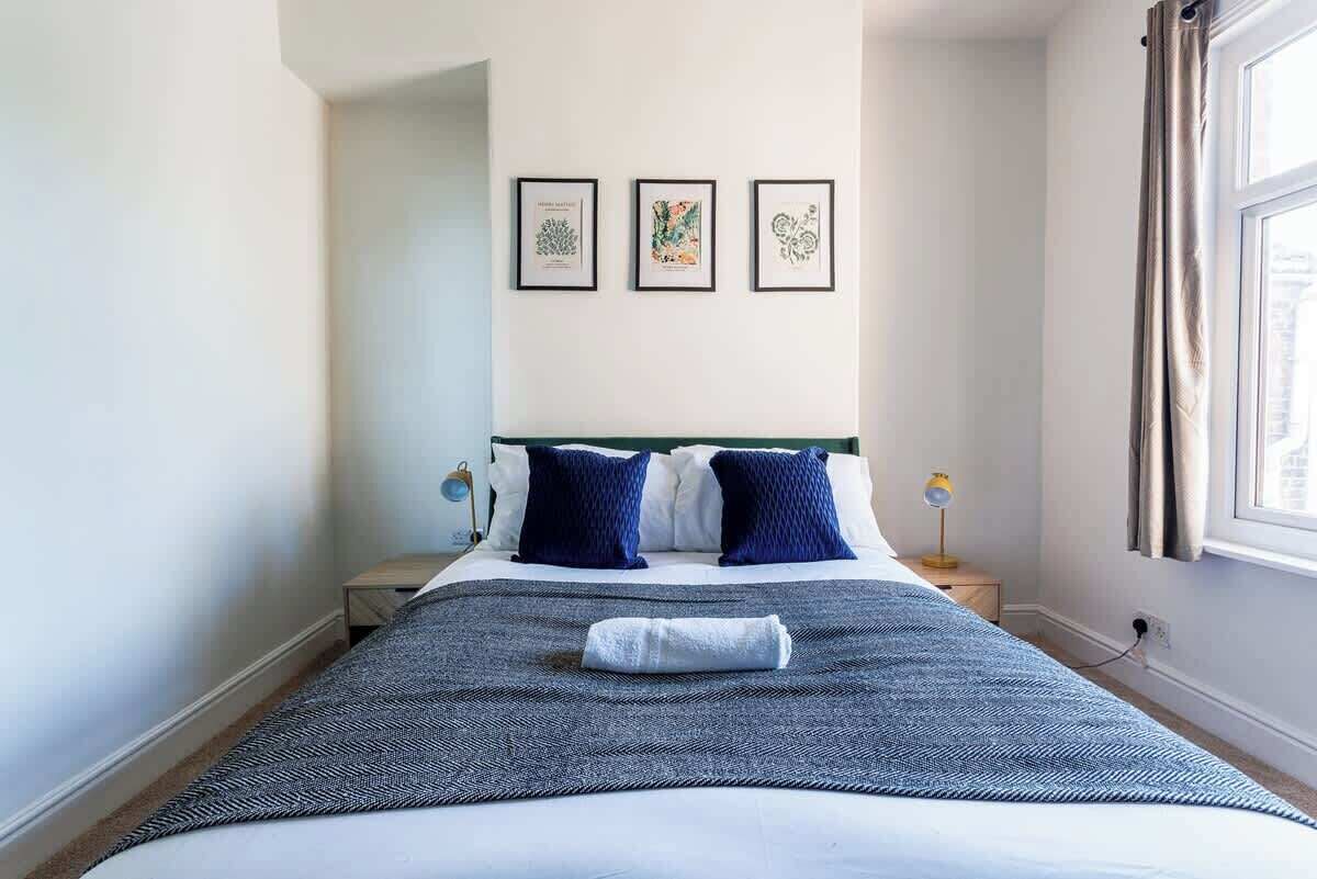 A neatly made bed with a blue and white patterned bedspread, two dark blue pillows, and a folded towel on the bed. There are three framed botanical prints on the wall above the bed.