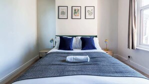 Una cama bien hecha con una colcha azul y blanca estampada, dos almohadas azul oscuro y una toalla doblada sobre la cama. Encima de la cama, hay tres láminas botánicas enmarcadas en la pared.