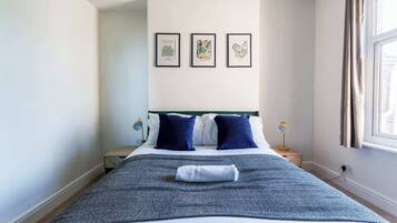 A neatly made bed with a blue and white patterned bedspread, two dark blue pillows, and a folded towel on the bed. There are three framed botanical prints on the wall above the bed.