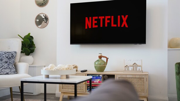 Un salon avec un téléviseur affichant le logo « NETFLIX », un canapé blanc, une table basse, une plante et un meuble en bois rempli de livres.
