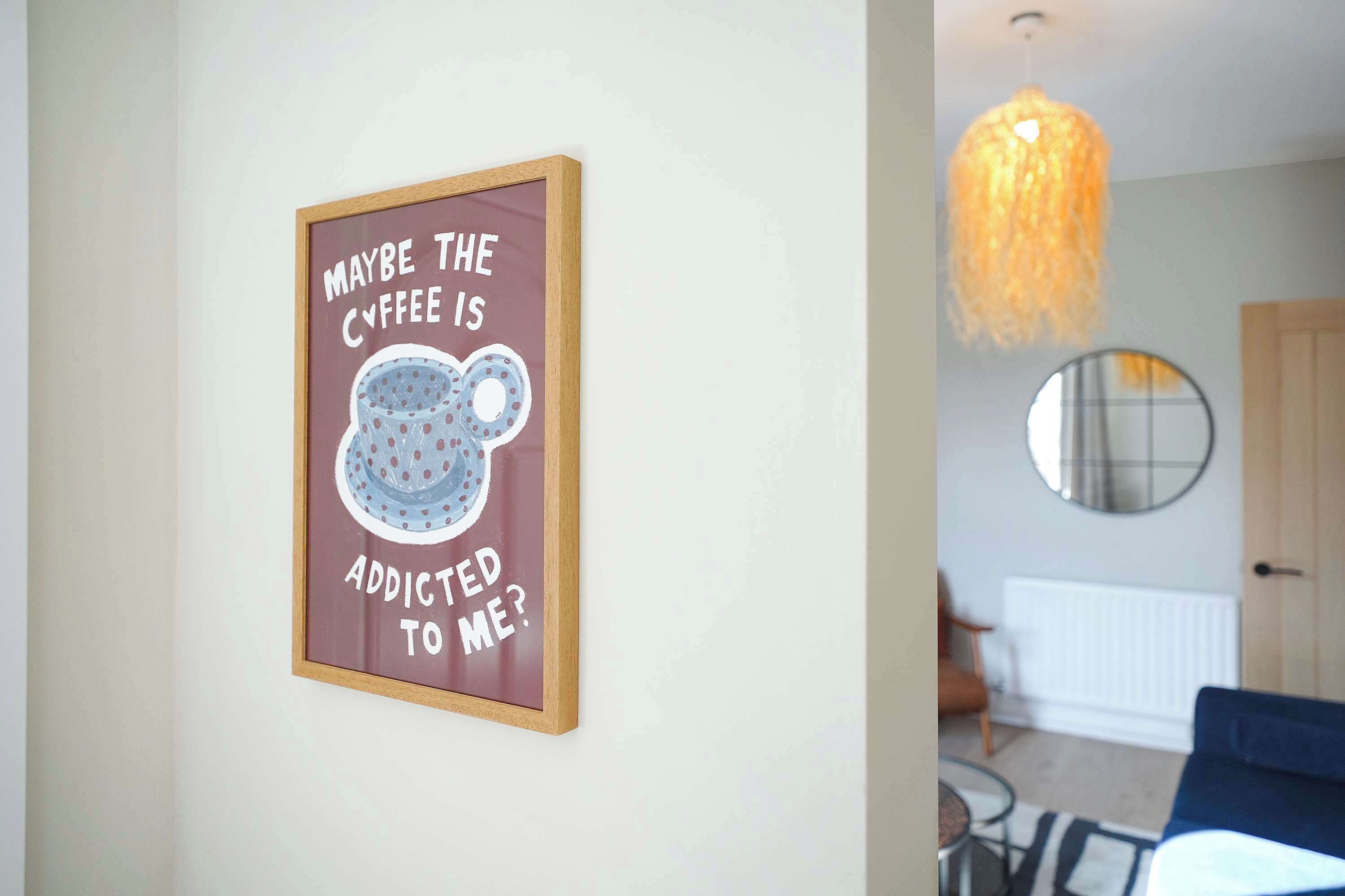 Un panneau encadré avec une tasse de café et du texte, un luminaire suspendu, un miroir rond et une porte dans une pièce.