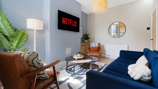 Un salon avec un canapé bleu, un fauteuil en cuir marron, une table basse ronde, un téléviseur affichant le logo « NETFLIX », une lampe, un miroir et une plante.