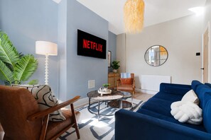 Ein Wohnzimmer mit einem blauen Sofa, einem braunen Ledersessel, einem runden Couchtisch, einem Fernseher mit dem Schriftzug 'NETFLIX', einer Lampe, einem Spiegel und einer Pflanze.