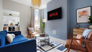 Een woonkamer met een blauwe bank, een bruine leren fauteuil, een glazen salontafel en een flatscreen-tv waarop 'NETFLIX' staat.