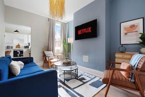 Ein Wohnzimmer mit einem blauen Sofa, einem braunen Ledersessel, einem Glastisch und einem Flachbildfernseher, der 'NETFLIX' zeigt.