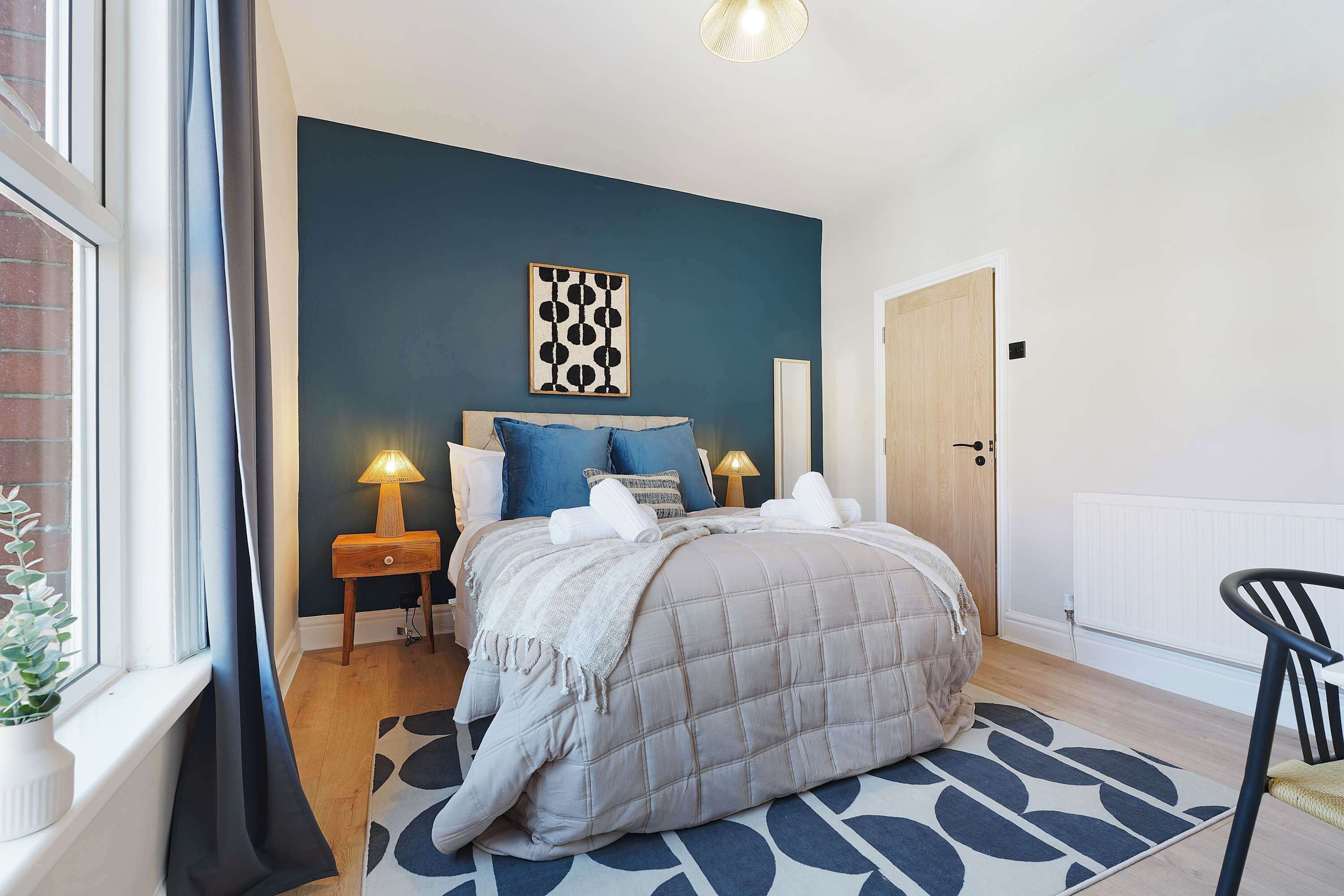 Une chambre à coucher comprenant un lit, des tables de chevet, une lampe, une chaise, un radiateur et une fenêtre avec des rideaux.