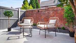 Twee rieten stoelen met kussens, een bakstenen muur en een potplant in een patio.