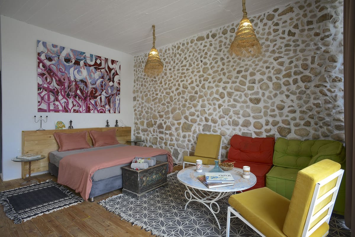 Un dormitorio con pared de piedra, una cama con ropa de cama rosa, una mesa redonda con un libro y una silla amarilla.